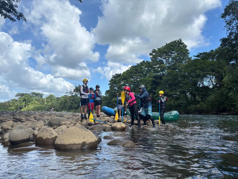 The image shows a group of people preparing for a rafting trip. They are standing on a rocky shore next to a river, wearing helmets and life vests. Some are holding paddles, and a raft is visible in the background. The sky is cloudy, and trees line the riverbank.